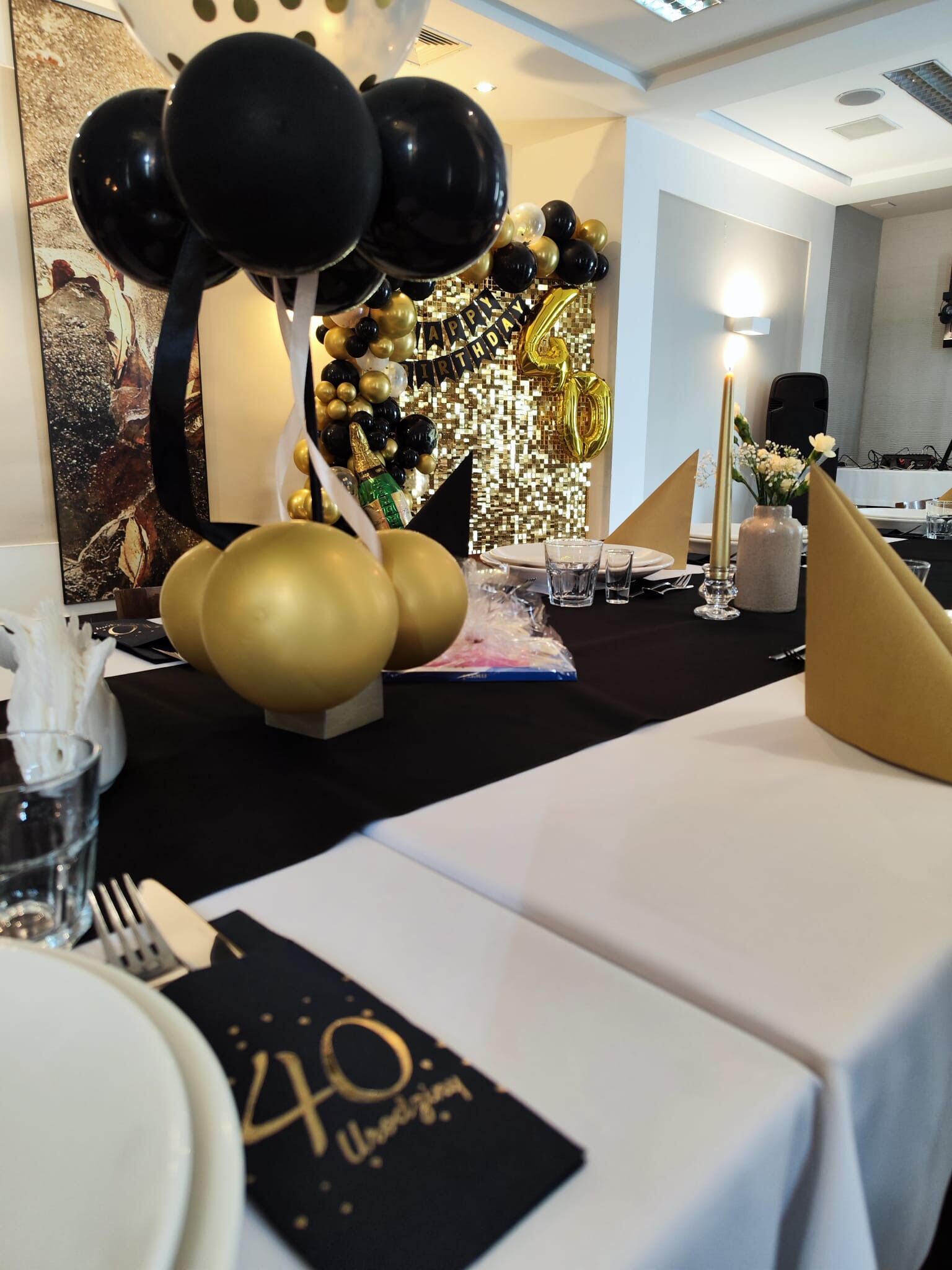Uma mesa posta para uma celebração, com balões, uma placa com os dizeres '40º Aniversário' e um painel decorativo.