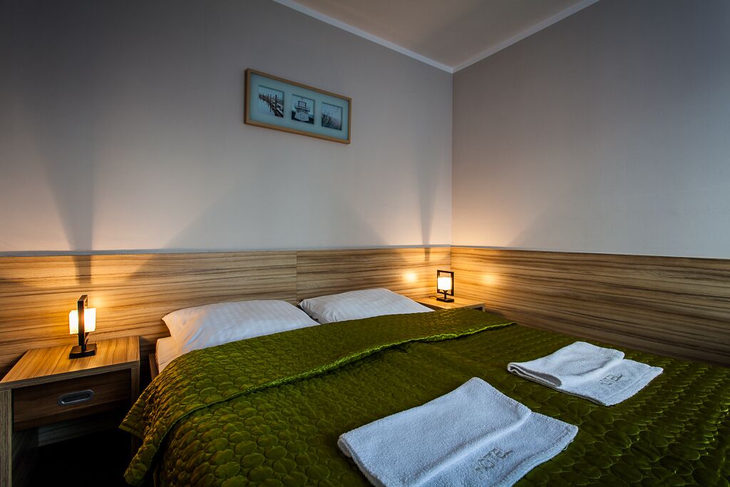 Habitación de hotel con cabecera de madera, colcha verde acolchada, dos almohadas blancas, mesita de noche con lámpara y un cuadro en la pared.
