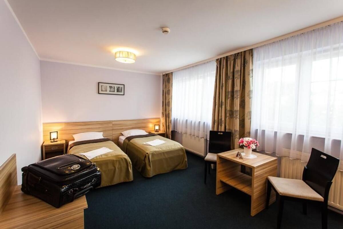 A hotel room with two single beds, a desk with a chair, a table with a vase of flowers, a suitcase, and a framed picture on the wall.