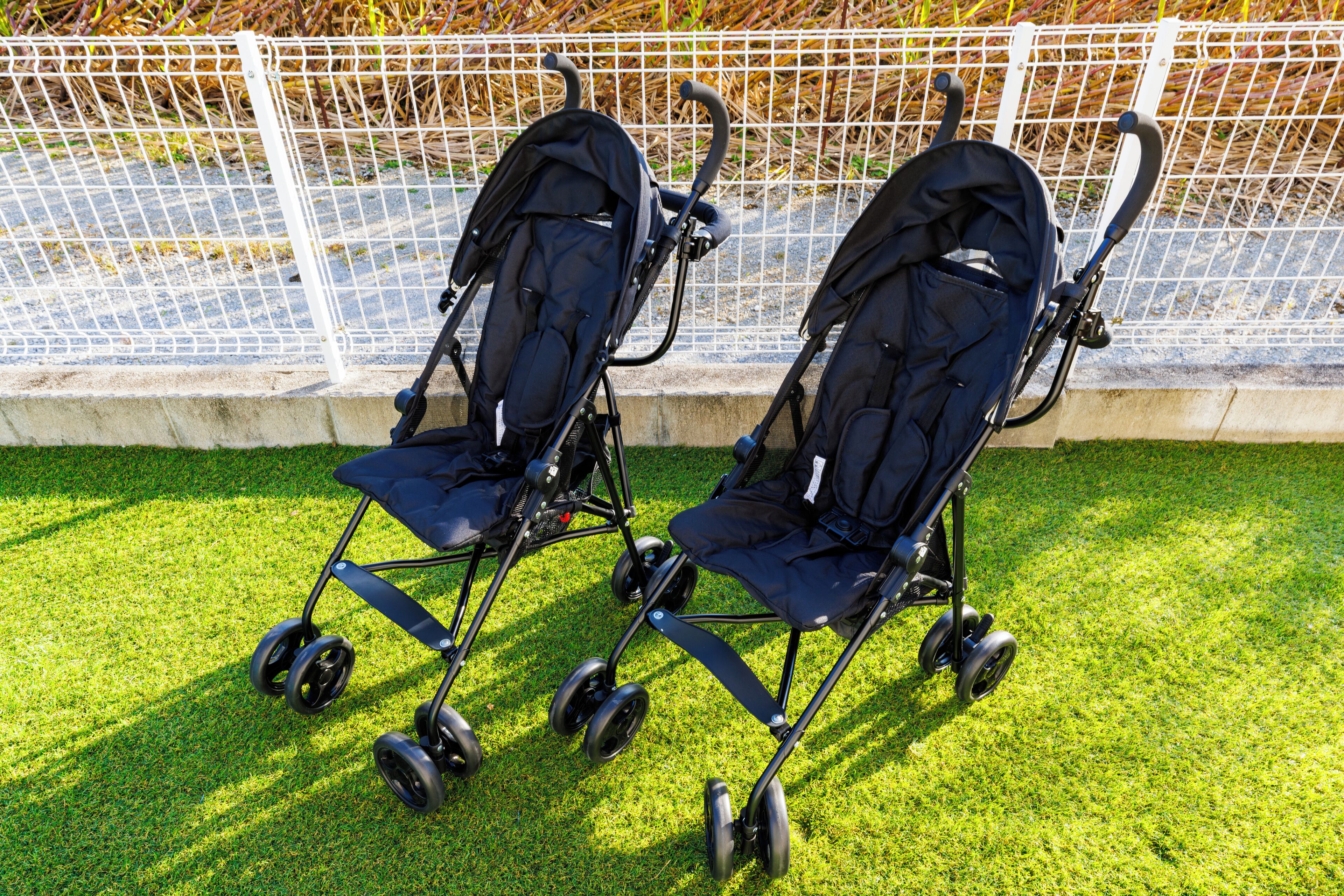 Two strollers parked on a grassy area with a white fence in the background.