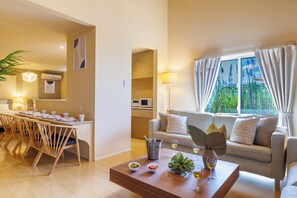 Een moderne woonkamer met een bank, een eettafel met stoelen, een salontafel en uitzicht op groen buiten het raam.