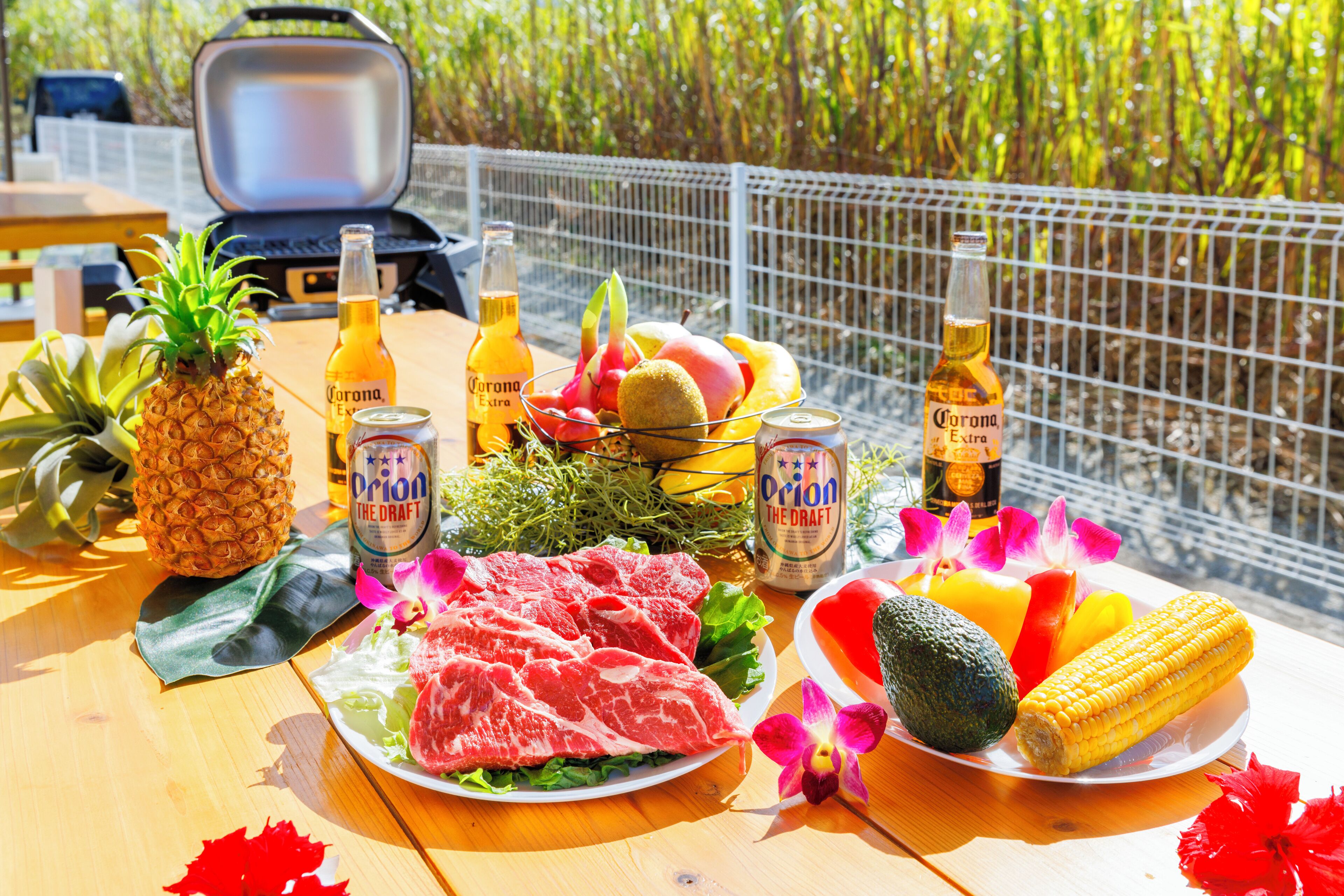 A table with food and drinks, including a pineapple, meat, and bottles of Corona Extra beer, set outdoors with a grill in the background.