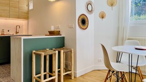 A modern kitchen with wooden cabinets, a white countertop, and a small island with stools. There are decorative wall hangings and a window with curtains. - Self-contained apartment in guest house - Near Paris (Meudon)