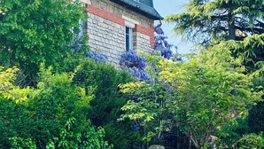 A two-story stone building with a gabled roof, surrounded by lush greenery and flowering plants. - Self-contained apartment in guest house - Near Paris (Meudon)