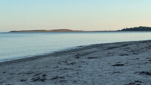 Una playa con cielo despejado, agua tranquila y colinas al fondo.
