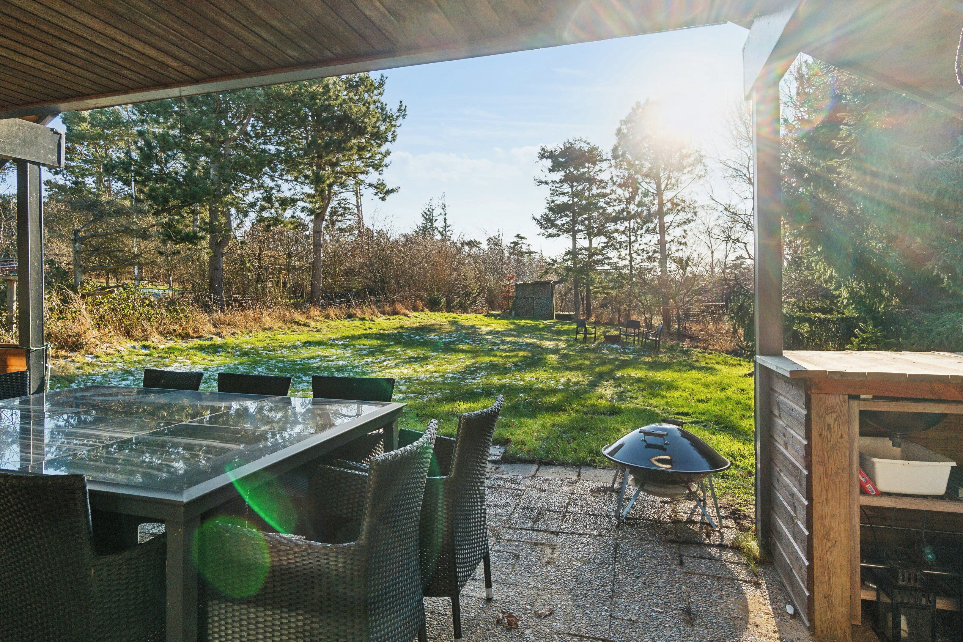 Een terras buiten met een tafel en stoelen met een glazen blad, een barbecue en een houten constructie.