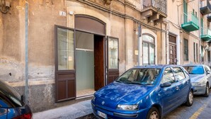 A blue car parked on a street with a building featuring a large arched door and a balcony. - GuestHost - Lovely 56 sqm apartment, for 2 people, located on the ground floor of a building (one step to access). The property is located in a central area, less than 10 minutes on foot from the station, in an area full of services and in a strategic po (Catania)