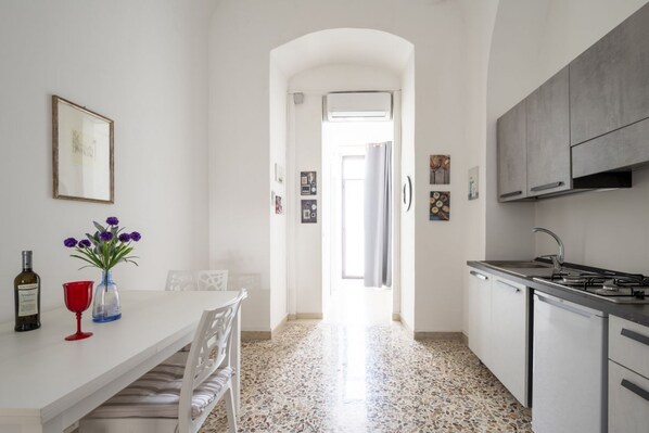 A kitchen with white cabinets, a countertop, and a stove. There is a dining area with a table and chairs, and a hallway leading to an open door.