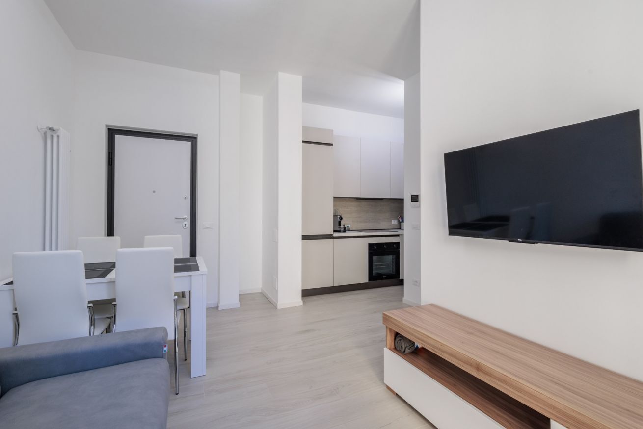 A modern living room with a flat-screen TV, a sofa, a dining table with chairs, and a kitchen area in the background.