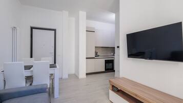 A modern living room with a flat-screen TV, a sofa, a dining table with chairs, and a kitchen area in the background.
