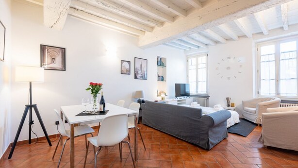 A room with a dining table, chairs, a sofa, a TV, and a clock on the wall. - Comfortable 55 sqm apartment in the heart of Parma. The accommodation, which can host up to 4 guests, is located on the first floor of a period building without lift. Please note that there is one step to access the building and stairs to reach the apart (Parma)