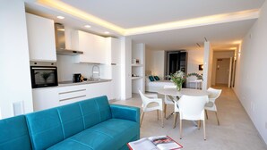 A modern living room with a teal sofa, a dining area with white chairs, and a kitchen with white cabinets and a built-in oven. - [Calammare Apartment N°5 Tramontana] - Seaview - Molfetta - Private Beach & Gym (Puglia)