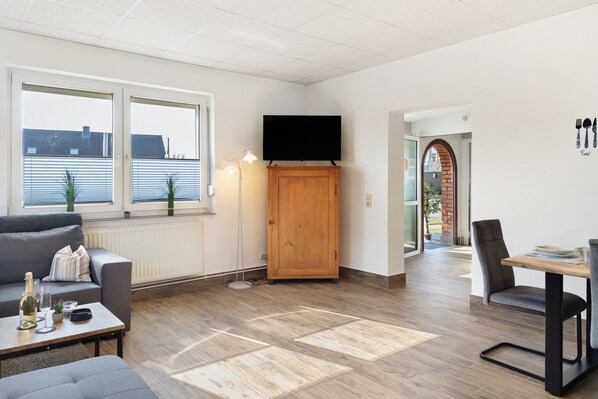 Una sala de estar moderna con un sofá, una mesa con una botella y vasos, un televisor sobre un mueble de madera, y un comedor con una silla y una mesa.