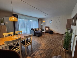 A modern living room with a dining area, wooden flooring, and a large window with sheer curtains. - Holiday Apartment 'Ursel' with Private Terrace, Garden, and Wi-Fi (Büsum)