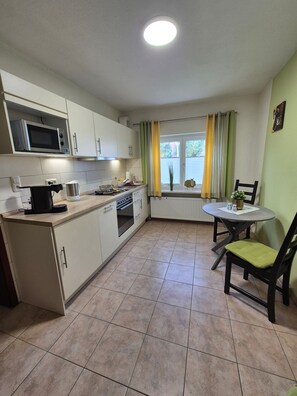 A compact kitchen with a microwave, stove, and sink, a dining table with chairs, and a window with curtains.