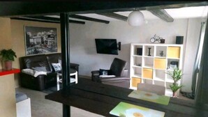 Un salon avec un canapé, un fauteuil et un téléviseur fixé au mur. On y trouve une bibliothèque remplie de divers objets et un vélo posé sur le dessus.