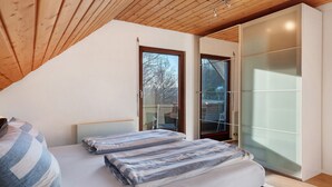 Une chambre avec un plafond en pente, un lit avec une literie à rayures, une porte-fenêtre coulissante et une armoire blanche.