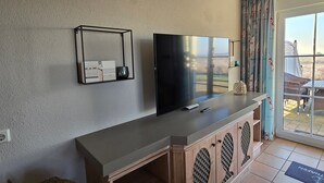 Ett modernt vardagsrum med en platt-TV, en liten hylla med en bok och ett ljus, och en skjutdörr i glas som leder ut till ett utomhusområde.