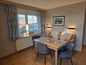 Un coin repas comprenant une table, des chaises, un radiateur, une fenêtre avec des rideaux et un tableau encadré accroché au mur.