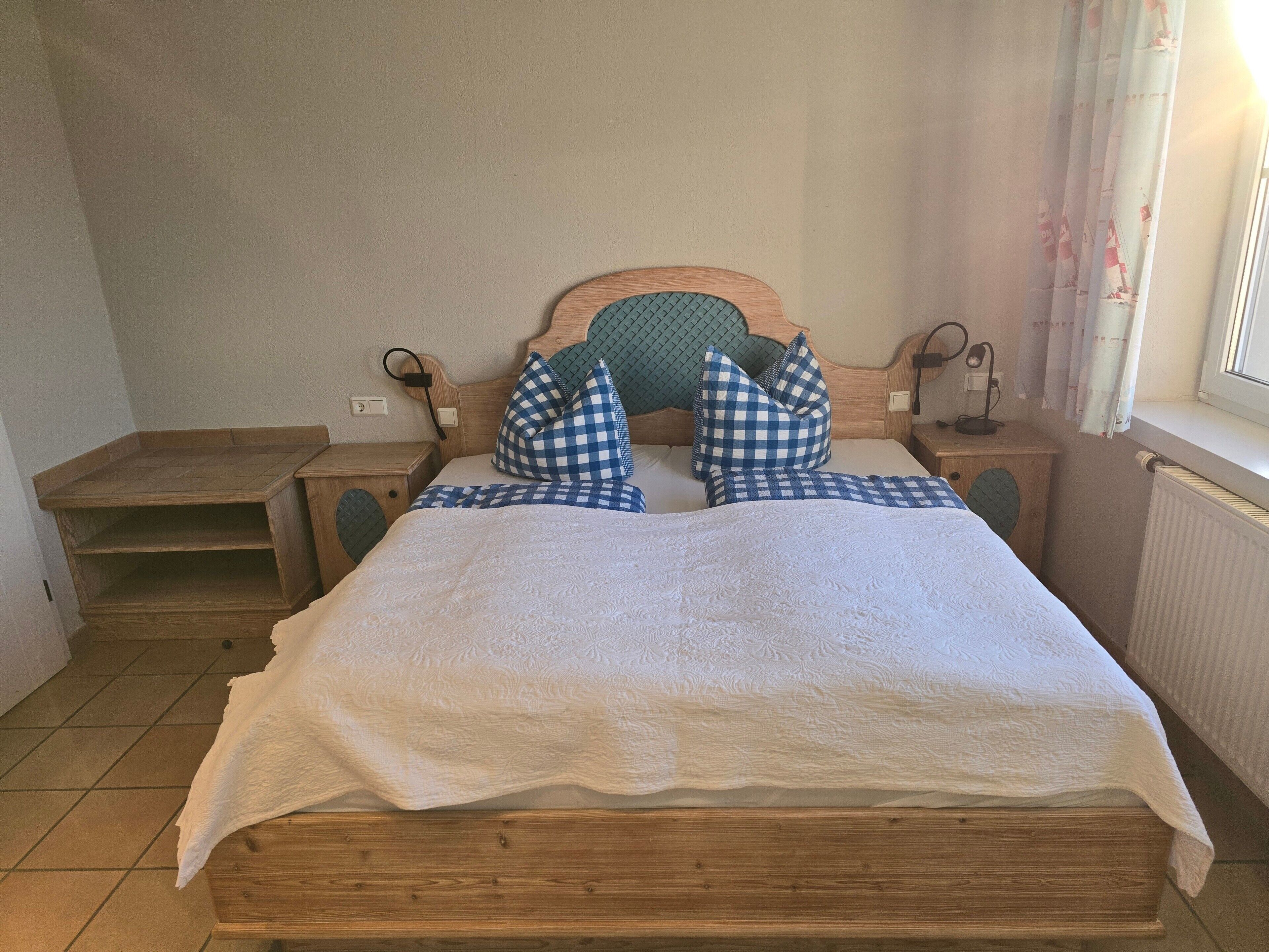 Un lit en bois avec une couvre-lit blanche et des oreillers à carreaux bleus, une table de chevet avec une lampe et une fenêtre avec des rideaux.