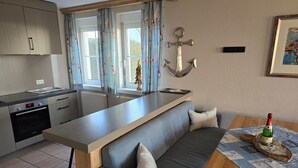 A modern kitchen with a built-in oven, a dining area with a wooden table and grey sofa, and a view of the outdoors through large windows. - Apartment 'Seeschwalbe' with Sea View, Shared Terrace and Wi-Fi (Mönchgut)