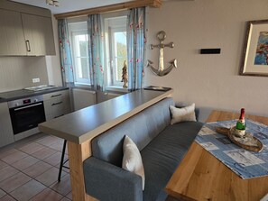 Een moderne keuken met een ingebouwde oven, een zithoek met een houten tafel en een grijze bank, en uitzicht op de omgeving door grote ramen.