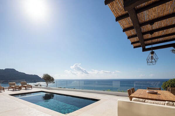 Un espace aménagé au bord de la piscine, avec une table et des chaises en bois, une balustrade en verre et une vue sur la mer.
