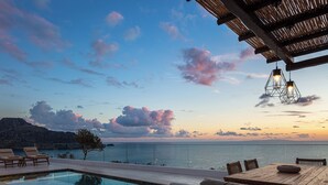 An infinity pool with lounge chairs and a wooden table, overlooking a body of water and a mountainous landscape.