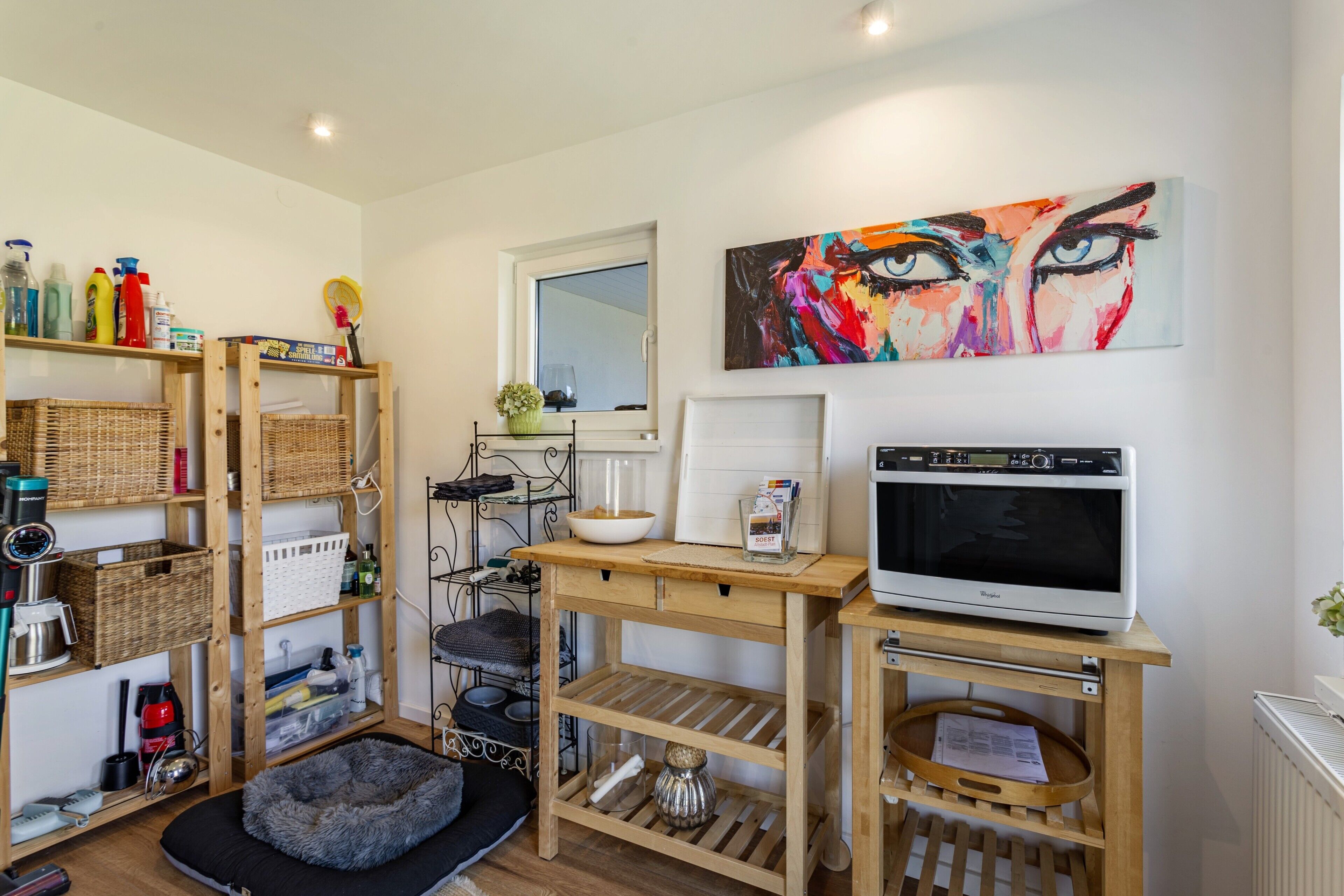 A room with a wooden shelf unit, a microwave, a painting, and a window.