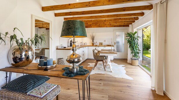 Un interior moderno con vigas de madera a la vista, un comedor con mesa y sillas, una lámpara y vistas por un ventanal grande.