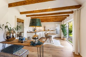 Ein modernes Interieur mit freiliegenden Holzbalken, einem Essbereich mit Tisch und Stühlen, einer Lampe und Blick durch ein großes Fenster.
