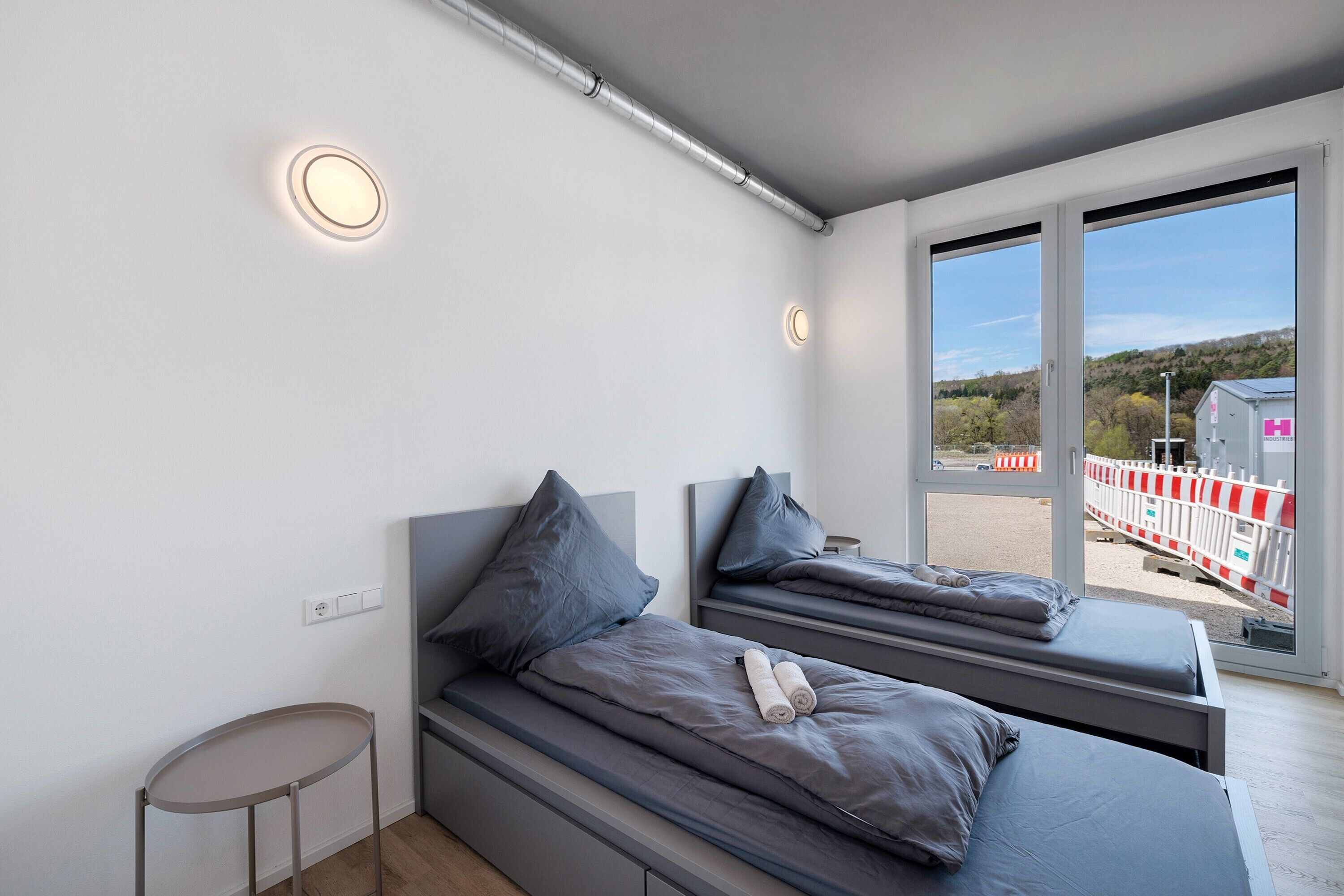 A modern bedroom with two beds, a small table, and a view of the outside.