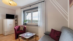 Ein Wohnzimmer mit einer Couch, einem roten Sessel, einem Couchtisch, einem Fernseher auf einer Kommode und einem Fenster mit Vorhängen.