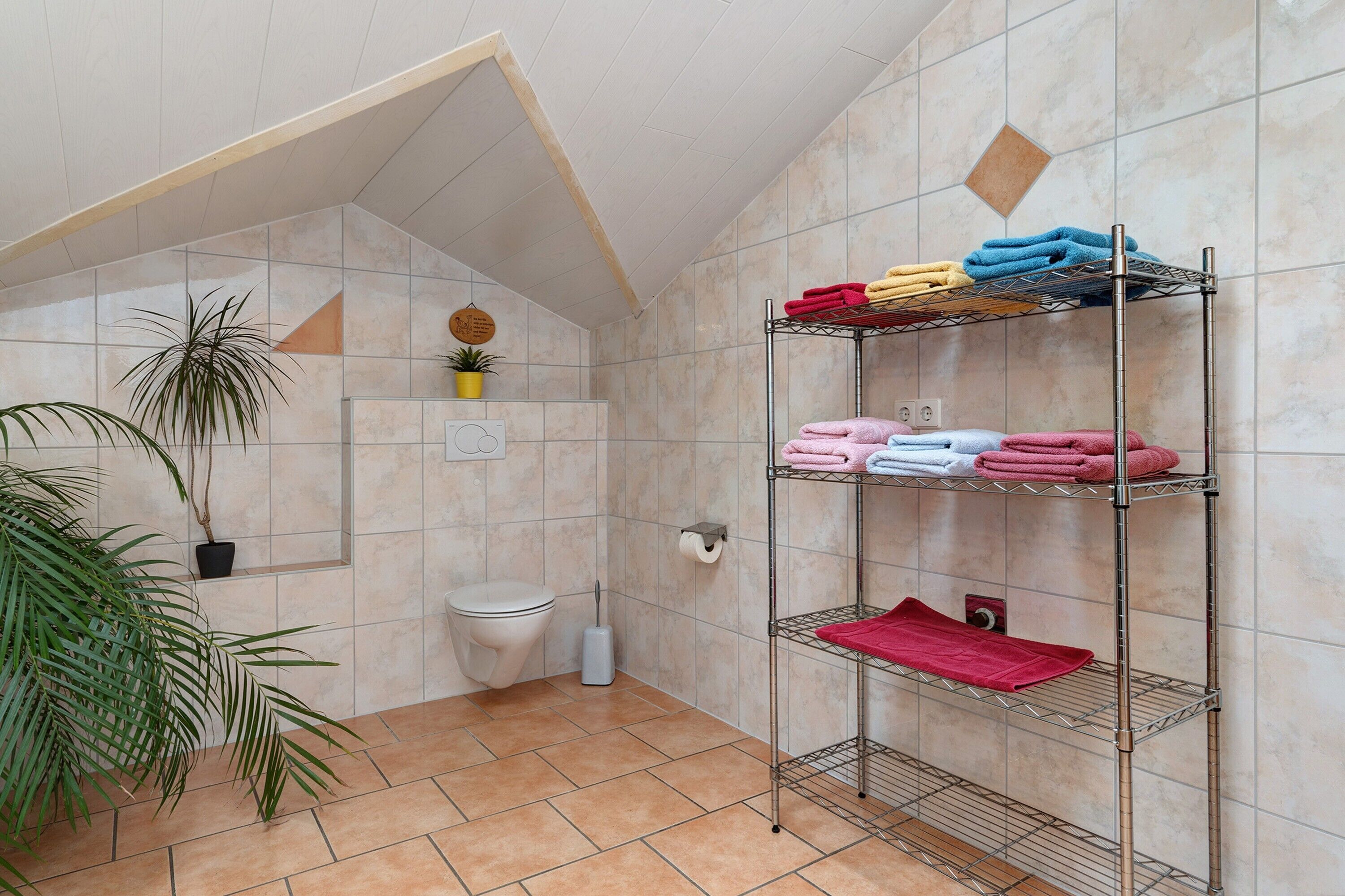 Un baño con inodoro, un estante de metal con toallas, una planta en maceta y piso de baldosas.