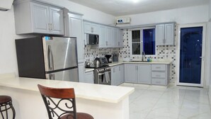 A modern kitchen with grey cabinets, a stainless steel refrigerator, a microwave, and a stove. There is a bar area with a white countertop and two chairs.
