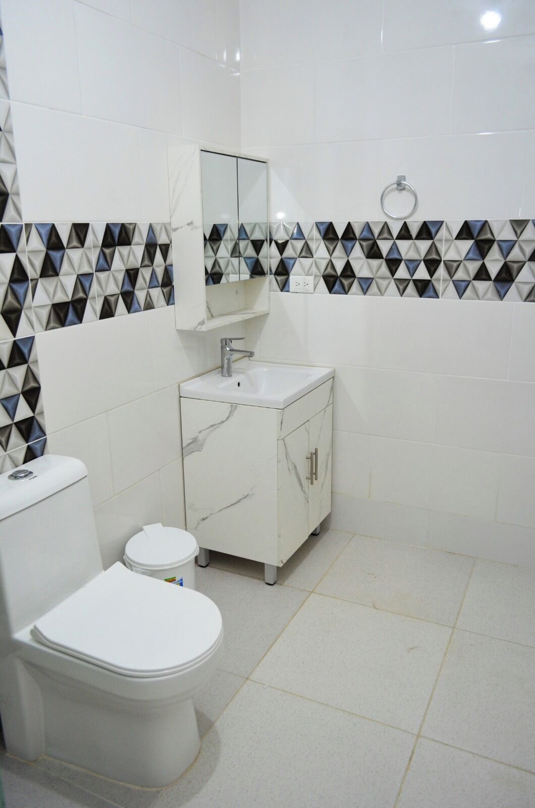 A bathroom with a toilet, sink, and cabinet, featuring white tiles and a geometric patterned backsplash.