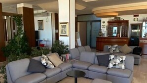 A modern living room with a large L-shaped sectional sofa, a small round coffee table, and a side table. There is a large mirror above the sofa and a wooden cabinet with glass doors in the background. - Hotel Spiaggia D'Oro (Mondolfo)