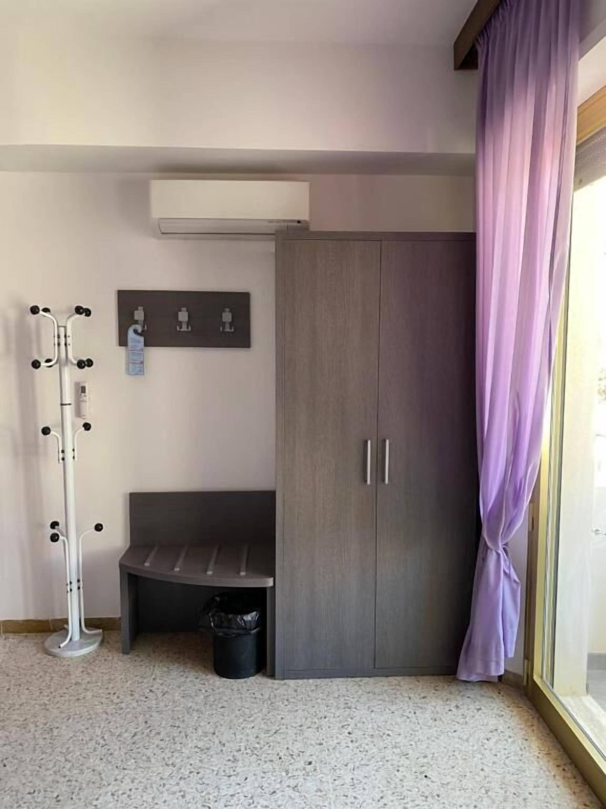 Una habitación con un armario gris, un banco, un perchero y una ventana con cortinas moradas.