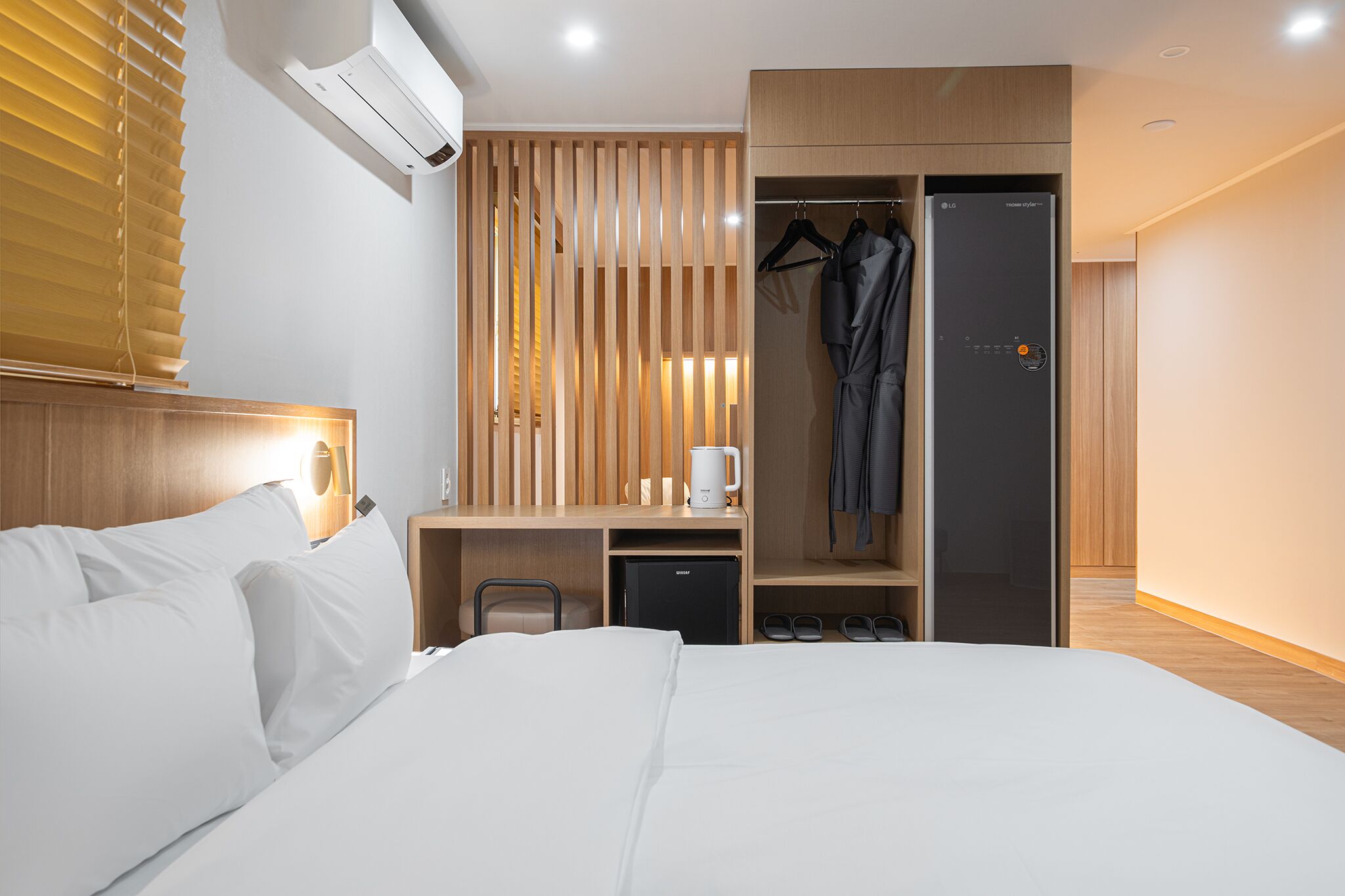 A hotel room with a bed, a desk with a coffee maker, a wardrobe, and a hanging coat.