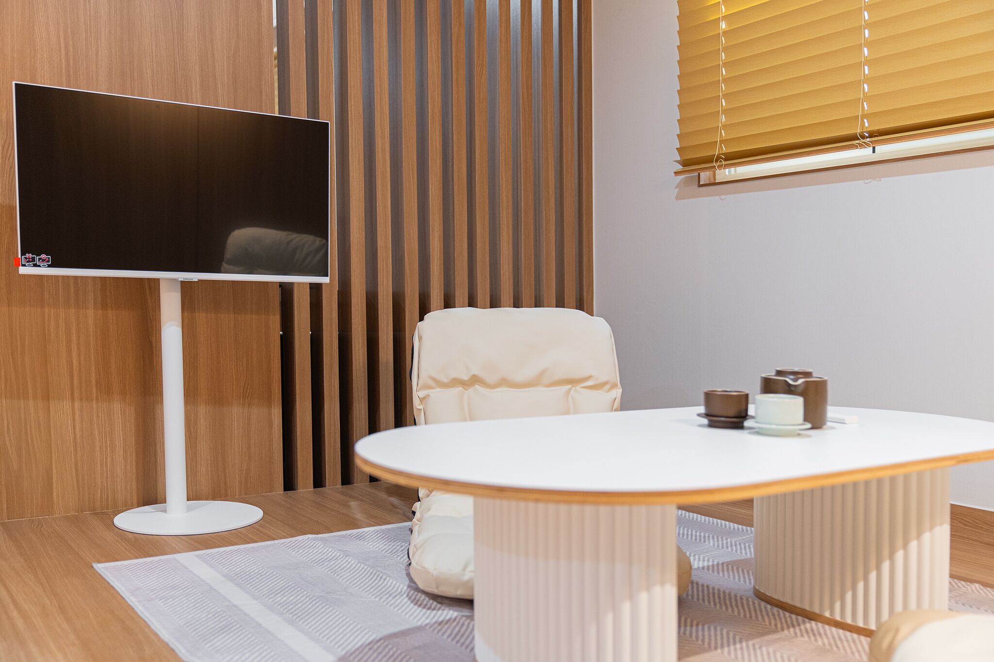 A modern room with a flat-screen TV mounted on a white stand, a round white table with a white chair, and a wooden slatted partition behind the TV.
