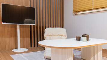A modern room with a flat-screen TV mounted on a white stand, a round white table with a white chair, and a wooden slatted partition behind the TV.