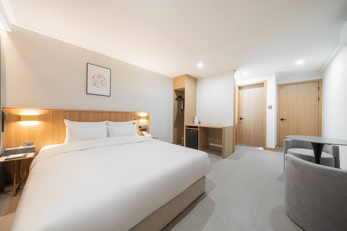 A hotel room with a large bed, bedside table, lamp, wardrobe, desk, and a small seating area.