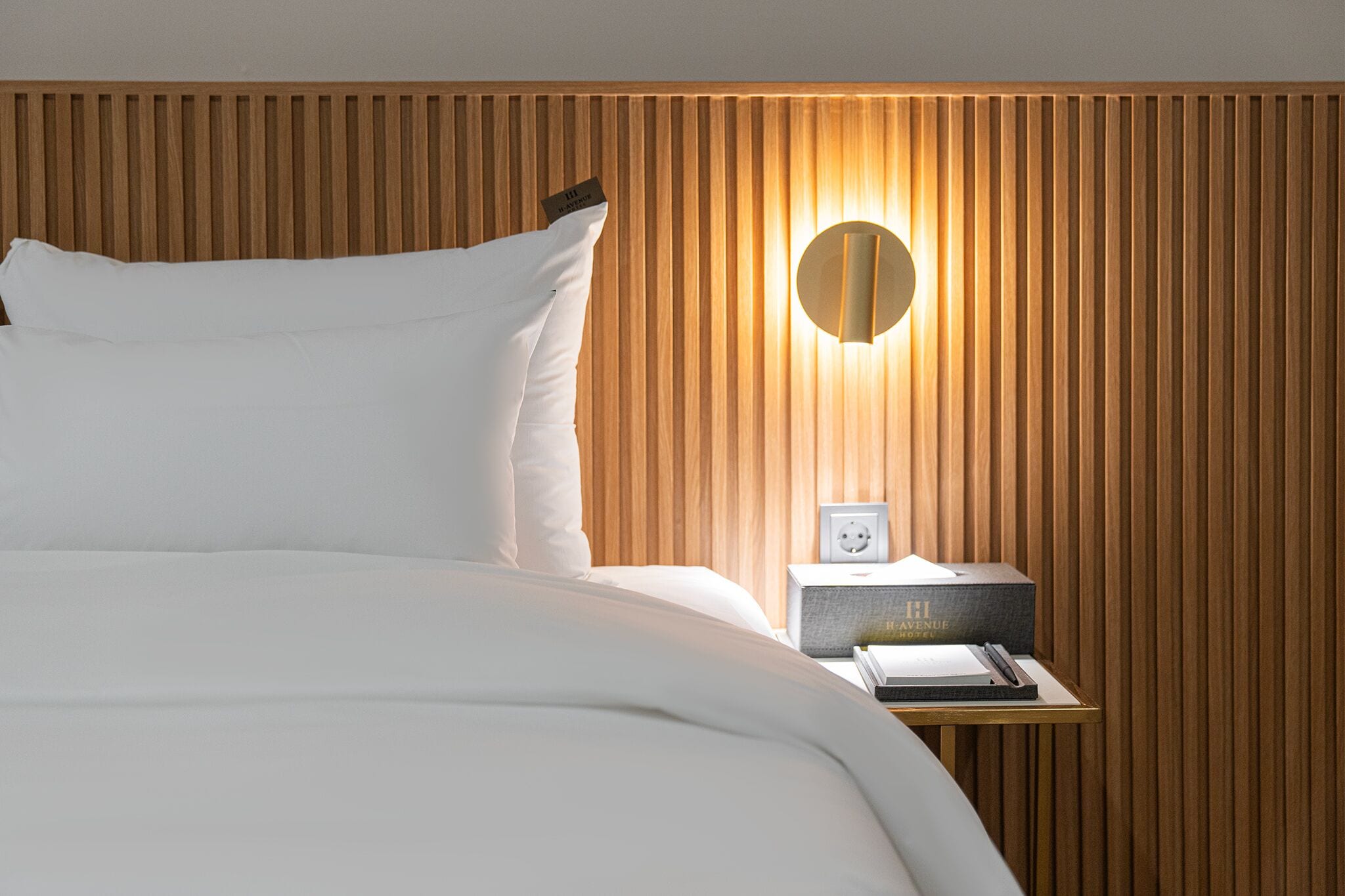 A neatly made bed with white linens, a wooden slatted headboard, a bedside table with a lamp, and a small box labeled "Haven Hotel."