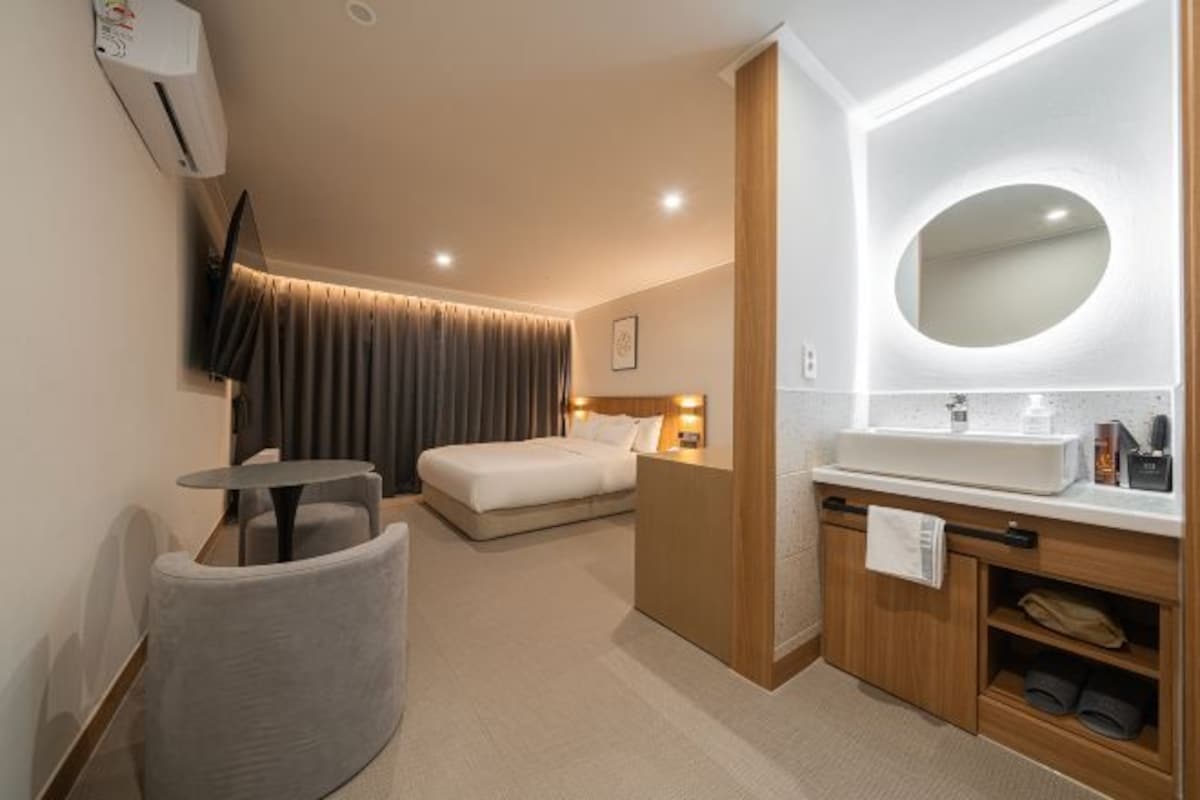 A hotel room with a bed, a small round table, a vanity with a mirror, and a wall-mounted air conditioner.