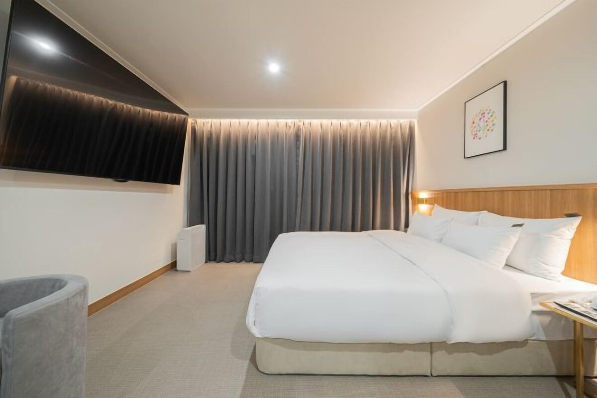 A hotel room with a bed, a TV mounted on the wall, a grey curtain, a white wall, and a framed picture.