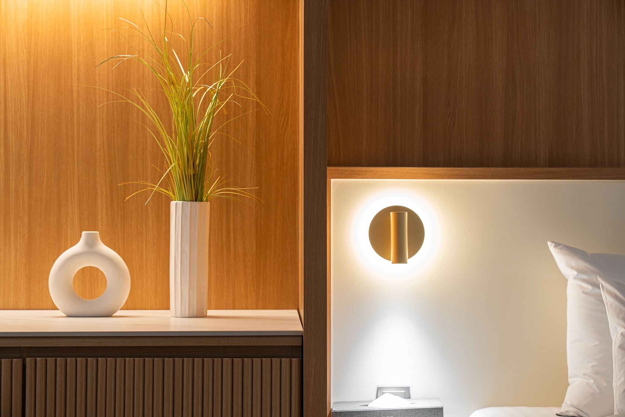 A modern hotel room with a wooden panel wall, a bedside table with a white vase and plant, and a wall-mounted light fixture.