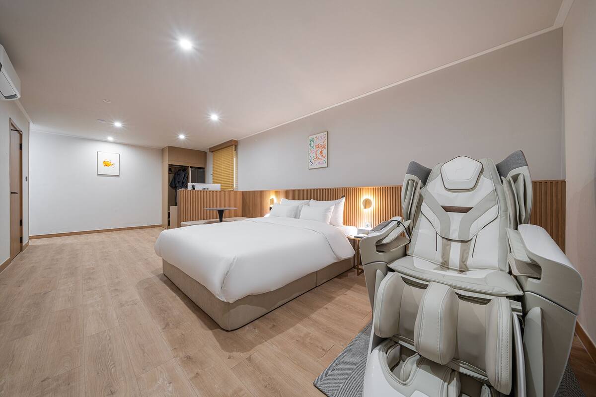 A hotel room with a large bed, a massage chair, a desk with a lamp, and a wardrobe.