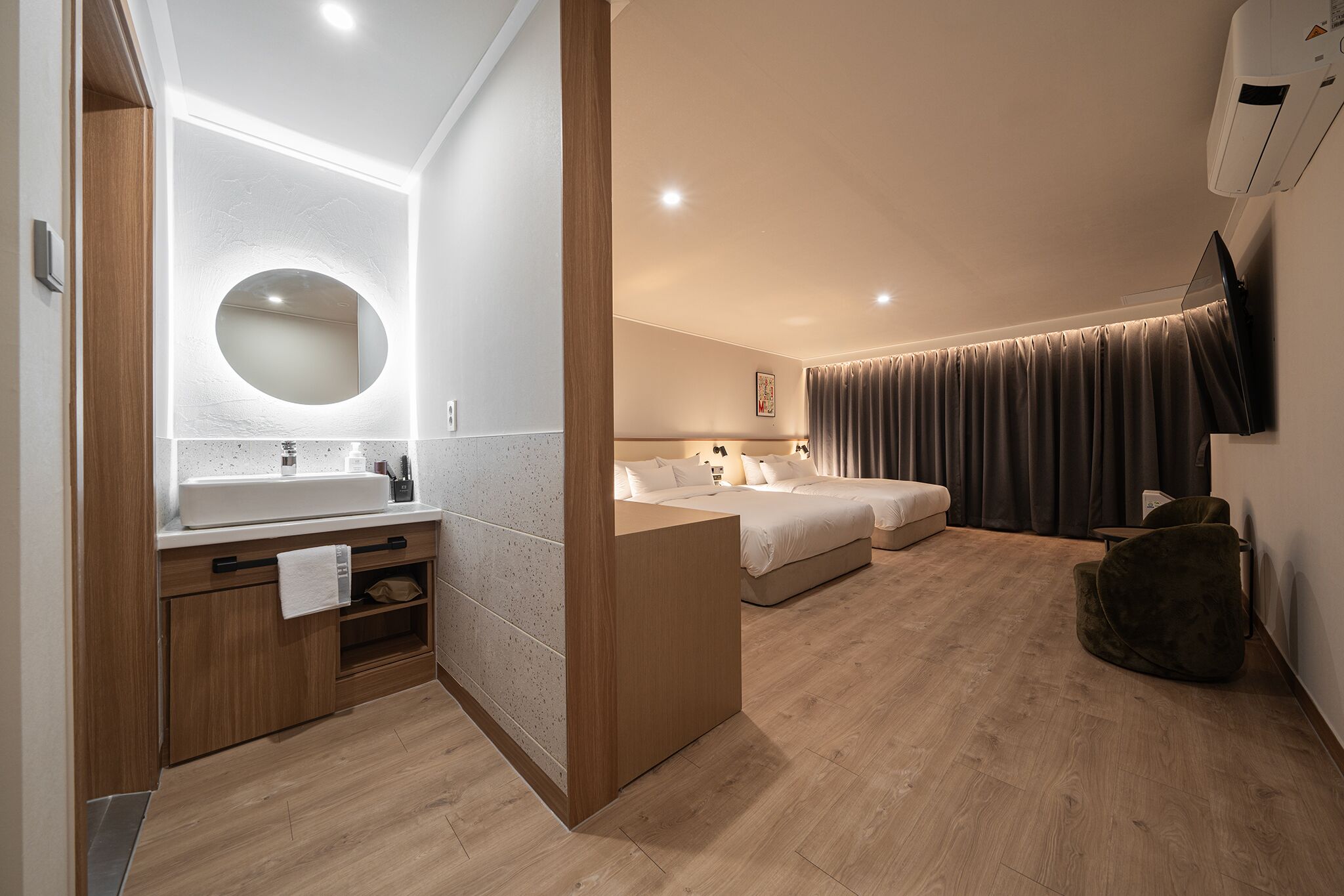 Una habitación de hotel moderna con piso de madera, un espejo redondo sobre el lavamanos, una cama con ropa blanca, un sillón de cuentas de color oscuro y un televisor de pantalla plana montado en la pared.