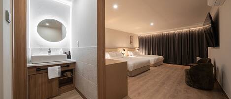 Una habitación de hotel moderna con piso de madera, un espejo redondo sobre el lavamanos, una cama con ropa blanca, un sillón de cuentas de color oscuro y un televisor de pantalla plana montado en la pared.