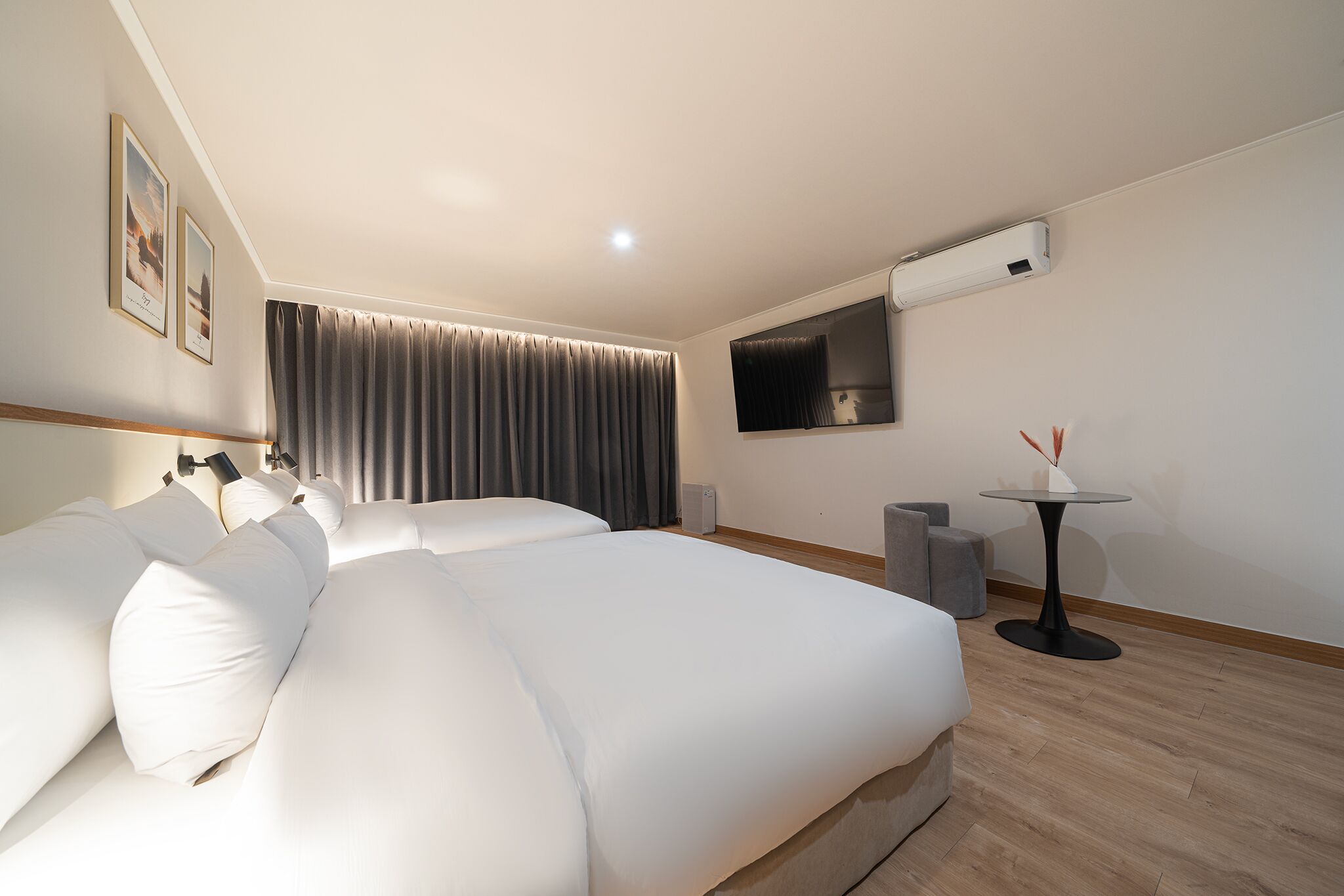 A hotel room with a large bed, a TV mounted on the wall, a small table, and a window with curtains.